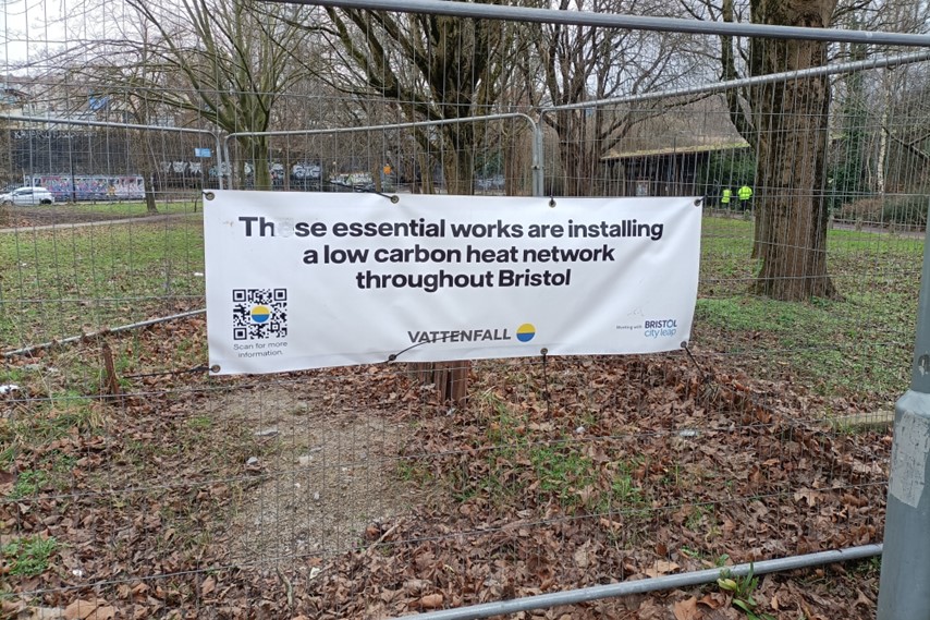 A sign hung on temporary fencing which reads "These essential works are installing a low carbon heat network throughout Bristol". Bedminster Green is visible in the background.